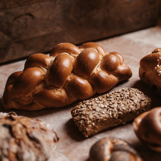 Ein Nahaufnahme verschiedener Brote auf einem Holztisch, einschließlich eines großen Challah-Laibs, eines Laibs mit Samen und anderer Brötchen.