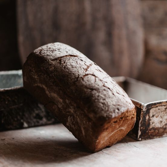 Ein Laib rustikales Brot liegt auf einem Holztisch, mit einer alten Metallbrotform dahinter. Das Brot hat eine strukturierte Kruste und sieht frisch gebacken aus.