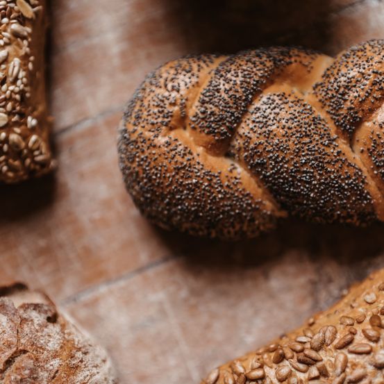 Drei Brote auf einer Holzoberfläche. Eines ist geflochten, ein anderes mit Samen bestreut und das dritte hat eine knusprige Kruste.