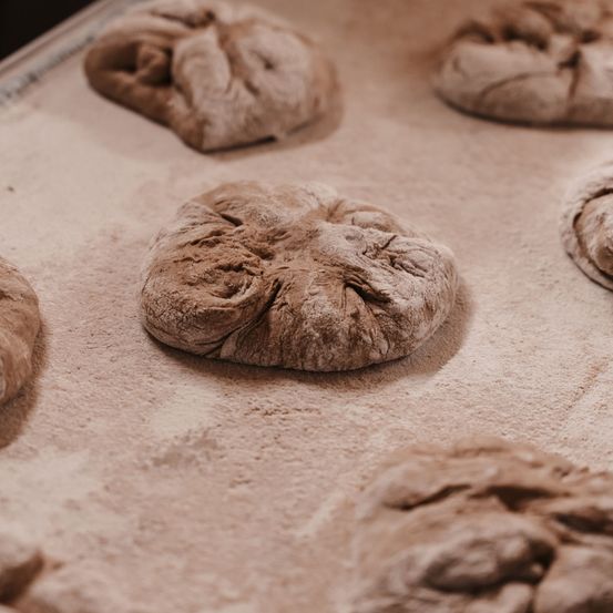 Mehrere ungebackene Brötchen liegen auf einem Tablett, mit Mehl bestreut, in einer Küchenumgebung.