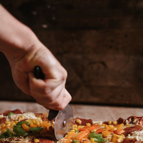 Eine Person schneidet eine Pizza mit verschiedenen Belägen, darunter Mais, Paprika und Pepperoni, mit einem Pizzaschneider.
