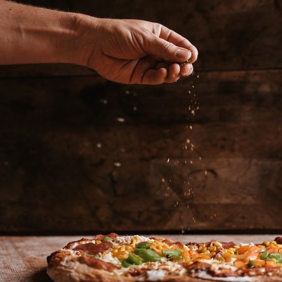 Eine Hand streut Pfeffer auf eine Pizza, die mit verschiedenen Gemüsesorten wie Mais und Paprika belegt ist und auf einer Holzoberfläche steht.