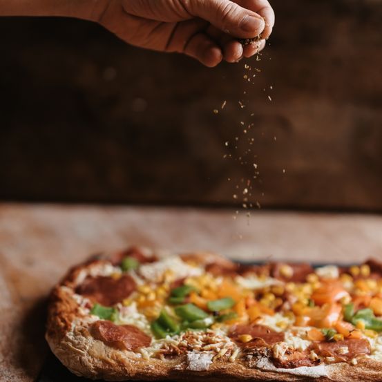 Eine Hand streut Pfeffer auf eine Pizza mit Toppings wie grünen Paprika, roten Paprika und Mais. Die Pizza ruht auf einem Holzschneidebrett.