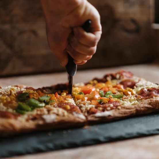 Eine Hand schneidet eine Pizza mit Gemüse und Käse auf einer Holzoberfläche.