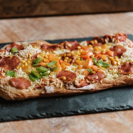 Eine Nahaufnahme einer Pizza mit Pepperoni, Mais, Paprika und Käse auf einem Holztisch.