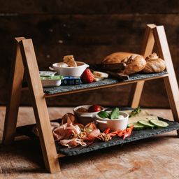 Ein hölzerner mehrstöckiger Tablett mit verschiedenen Speisen darauf. Die oberste Schicht hat eine Schüssel, eine Erdbeere und einen kleinen Teller. Die mittlere Schicht hat Brotscheiben und die untere Schicht hat Fleisch, Gemüse und Kräuter.
