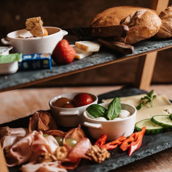 Ein hölzerner Tisch zeigt ein mehrstöckiges Tablett mit verschiedenen Speisen. Die oberste Etage besteht aus Brot, Käse und einer kleinen Schüssel mit Obst. Die untere Etage umfasst eine Schüssel mit Basilikum und Käse, ein Fleischtablett und Gemüse.