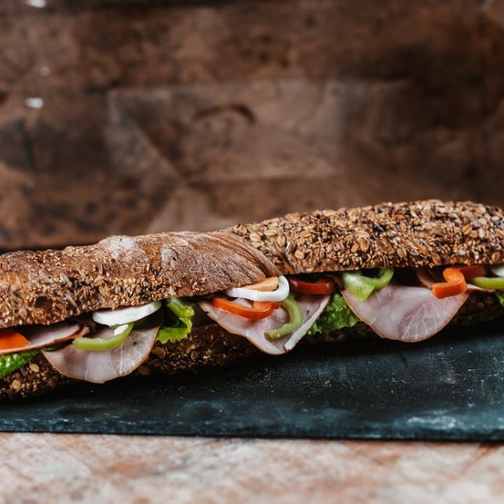 Ein langes Sandwich mit Körnern auf dem Brot, gefüllt mit Schinken, grünen Paprika, Tomaten und Zwiebeln. Es steht auf einem schwarzen Schieferteller vor einem hölzernen Hintergrund.