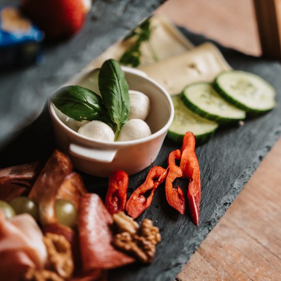Eine Charcuterie-Platte mit geschnittenen Gurken, roten Paprikaschoten und Mozzarella-Kugeln mit Basilikumblättern. Verschiedene Fleischsorten und Nüsse sind ebenfalls auf der Platte arrangiert.