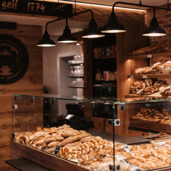 Eine Bäckerei mit Holzregalen zeigt verschiedene Brote, Gebäck und Brötchen. Oben hängen schwarze Lampen von der Decke. An der Wand befindet sich ein Logo und das Datum 1774.