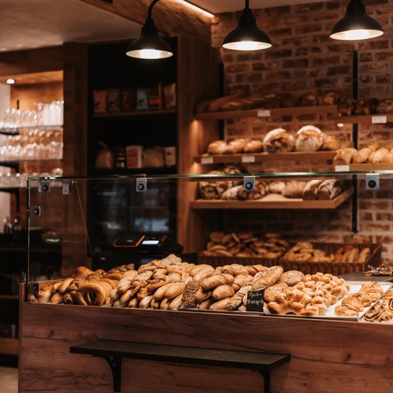 Eine Bäckerei gefüllt mit einer Vielzahl von Brot und Gebäck unter warmer Beleuchtung. Regale mit verschiedenen Brotsorten säumen die Ziegelwand. Eine Vitrine mit weiteren Broten und Gebäck.