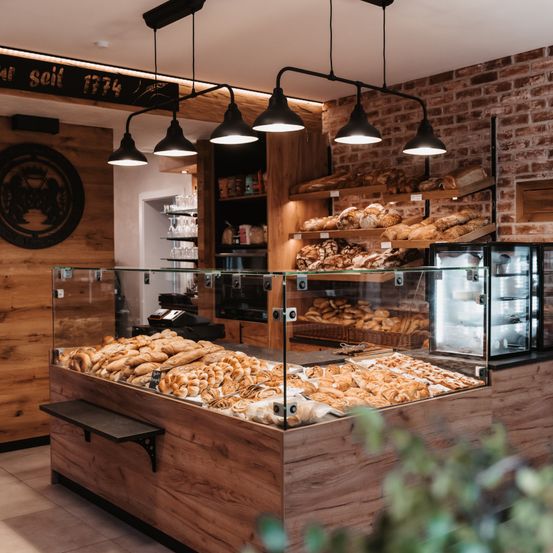 Ein Innenansicht einer Bäckerei mit Holzregalen und einer Ziegelwand, gefüllt mit verschiedenen Broten, Gebäck und Croissants, beleuchtet von hängenden Lampen.
