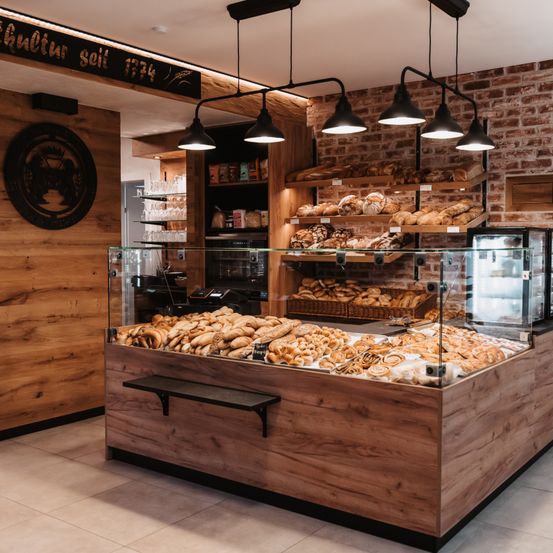 Das Innere einer Bäckerei mit Holzwänden, Ziegelakzenten und einer Glasvitrine, gefüllt mit verschiedenen Broten und Gebäck. Hinter der Theke befinden sich Regale mit weiteren Backwaren, und mehrere Lampen hängen von der Decke.