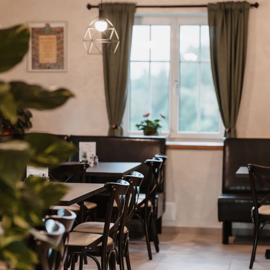 Ein Restaurant mit Tischen und Stühlen, einem Fenster mit Vorhängen und einer Topfpflanze auf der Fensterbank. An der Wand hängen ein Bilderrahmen und eine Hängelampe.