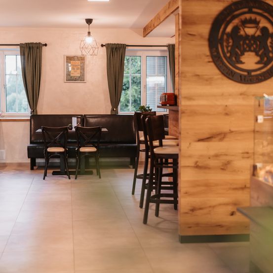 Ein leeres Restaurant mit einer Holztheke, einer Bank, Stühlen und einem Tisch mit Topfpflanzen. Der Raum hat eine Pendelleuchte, einen Bilderrahmen und Fenster mit Vorhängen.