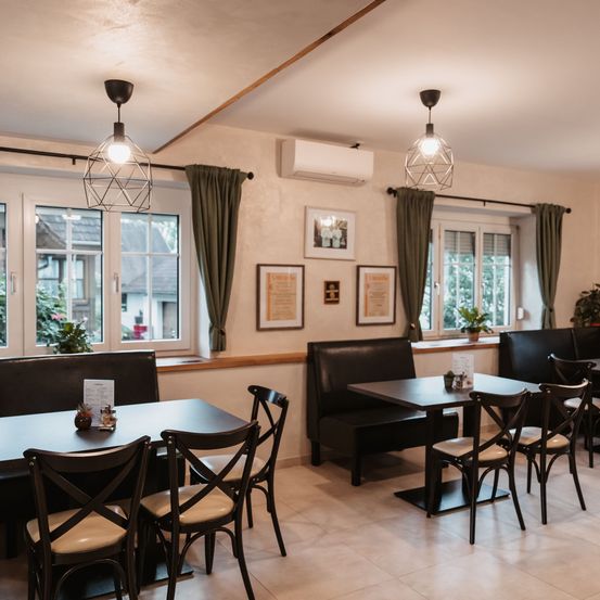 Ein Innenraum eines Restaurants mit weißen Wänden und beigen Böden. Es gibt einen langen Tisch mit Stühlen und eine Bank mit einem Topf auf dem.