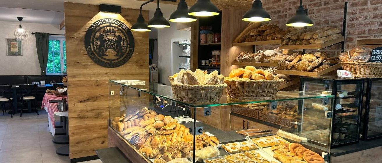 Eine Bäckerei mit Holzwänden hat eine Vitrine voller Brote, Croissants und anderen Gebäck. Der Innenraum wird von Pendelleuchten beleuchtet. Ein Schild an der Wand steht 'Bäckereimeister seit 1774'.