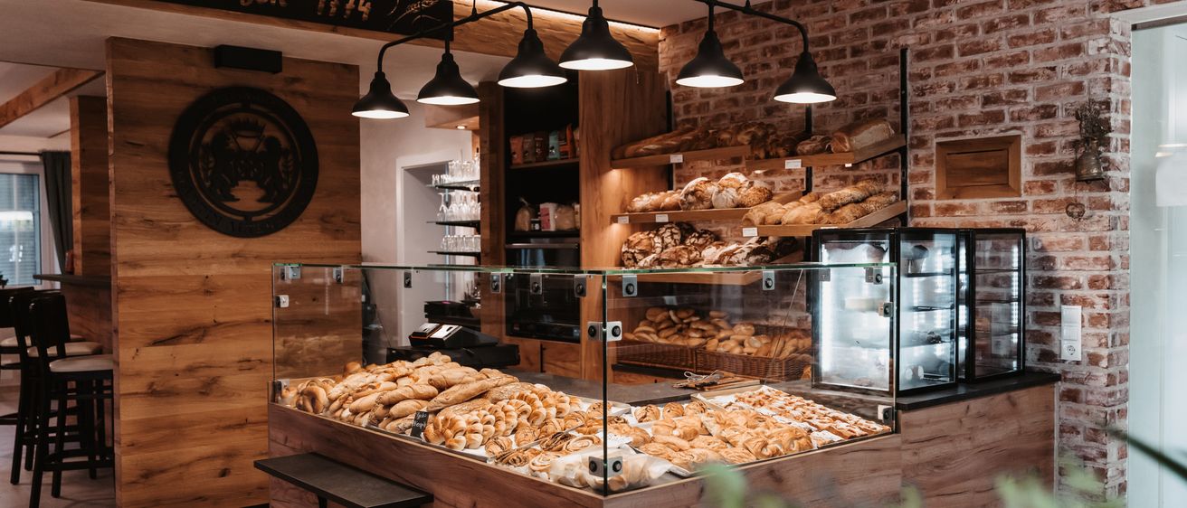 Eine Bäckerei mit einer Ziegelsteinwand und Holzregalen, die verschiedene Brote und Gebäck präsentieren. Eine Glasvitrine hält eine Auswahl an Brot bereit.
