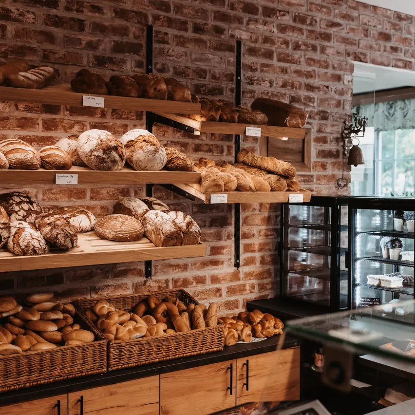 Eine Bäckerei mit einer Ziegelsteinwand zeigt verschiedene Brotsorten auf Regalen und in Körben. Ein Glasvitrine präsentiert Gebäck und Desserts.