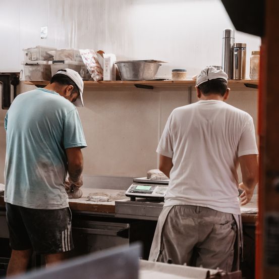 Zwei Männer arbeiten in einer Küche, einer trägt ein blaues Shirt und der andere ein weißes. Sie stehen vor einem Tisch mit einer Waage und verschiedenen Gegenständen in den Regalen.