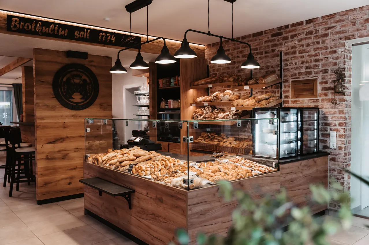 Das Innere einer Bäckerei mit Glasregalen, die eine Auswahl an Broten präsentieren, beleuchtet durch hängende Lampen, Holzwände und Ziegelmauerwerk.