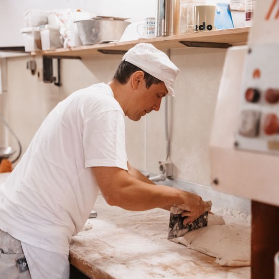 Ein Mann in einer weißen Kochmütze und einem weißen Kochschürze arbeitet mit einem Messer in einer Küche an Teig. Verschiedene Küchenutensilien und Behälter sind auf den Regalen zu sehen.