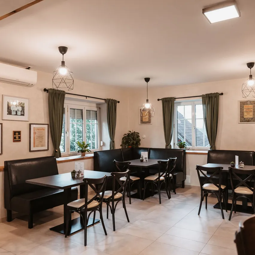 Ein gemütliches Restaurant mit modernen Möbeln, dunklen Ledersitzen und Holztischen. Der Raum hat weiße Wände, Deckenlampen und große Fenster mit grünen Vorhängen.