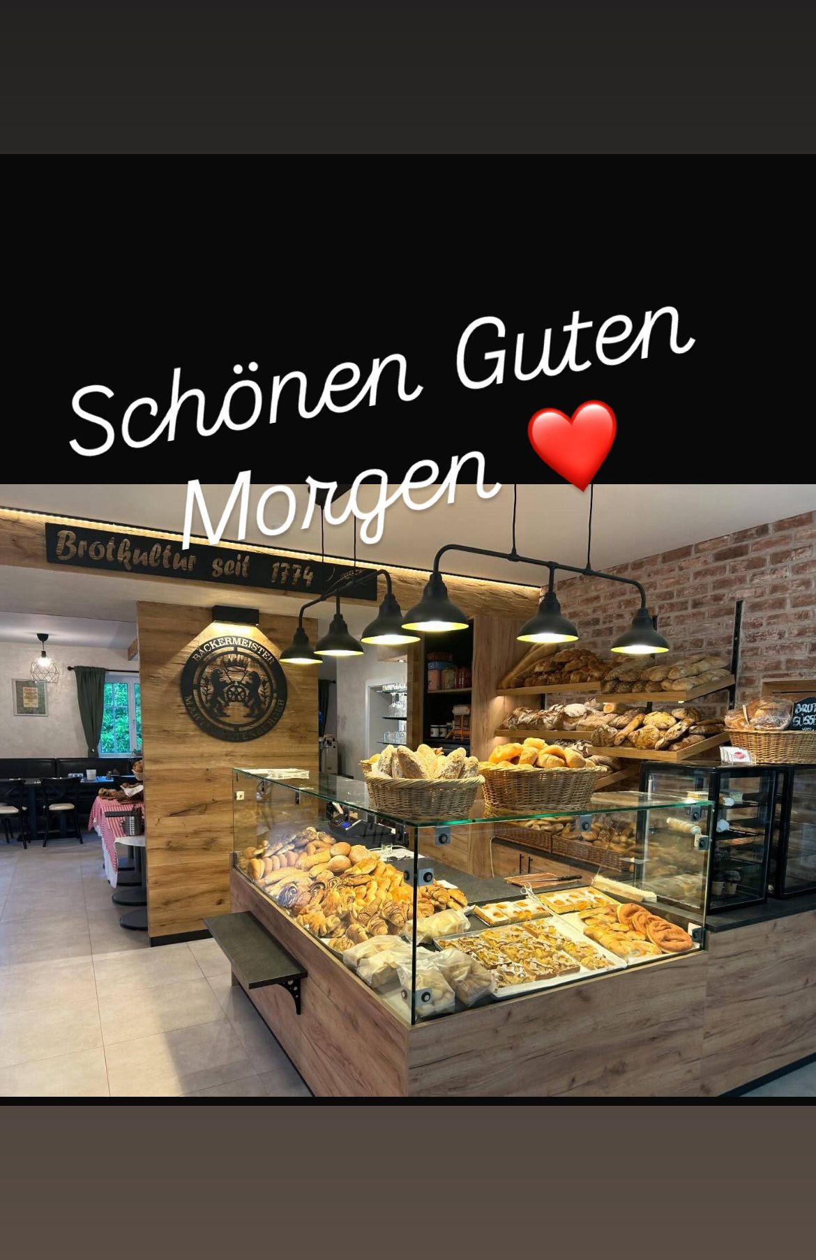 Ein Bäckerei-Interieur mit einer Glasvitrine voller Brot, Gebäck und anderen Backwaren. Der Tresen ist aus Holz und es hängen Lampen darüber. Die Worte 'Schönen Guten Morgen' sind oben angebracht.