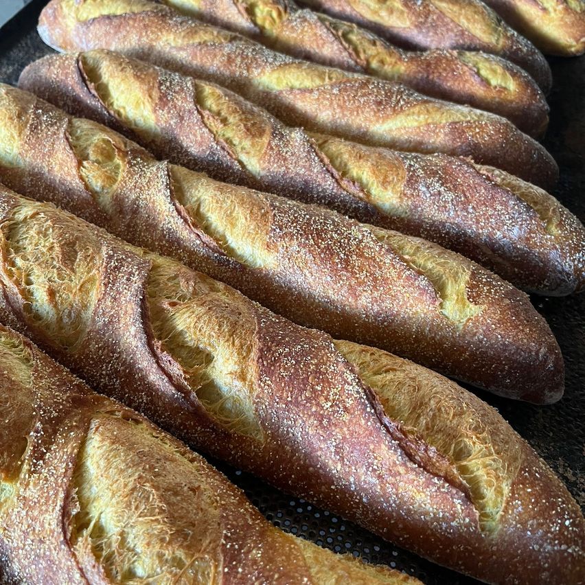 Ein Nahaufnahme von mehreren frisch gebackenen Baguettes, mit Mehl bestreut, auf einer schwarzen Oberfläche. Die Brote sind goldbraun mit einer leicht knusprigen Kruste.