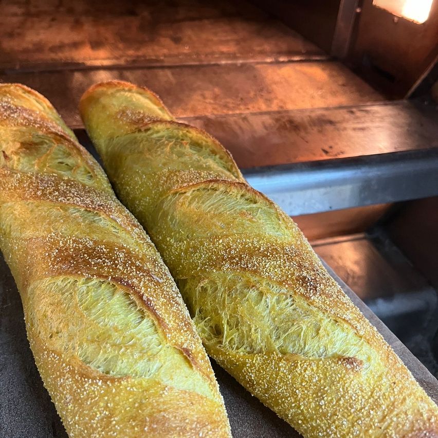 Zwei goldbraune Baguettes sind frisch gebacken und liegen auf einer schwarzen Oberfläche in einem Ofen mit einem warmen Licht darüber.