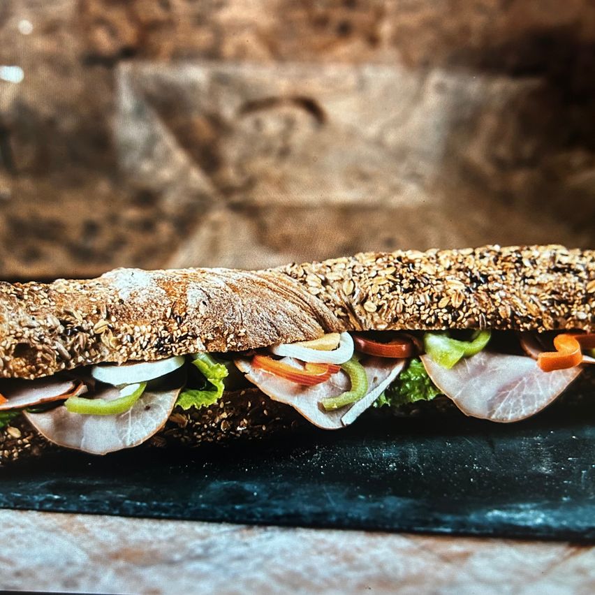 Ein großes Sandwich mit Schinken, Salat, Zwiebeln, Paprika und Karotten liegt auf einem schwarzen Schieferbrett.