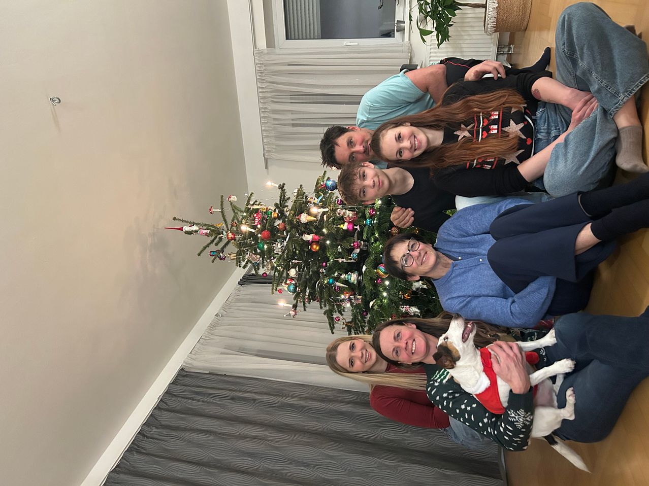 Eine fünfköpfige Familie posiert vor einem geschmückten Weihnachtsbaum, wobei eine Frau einen Hund hält. Sie tragen alle festliche Kleidung. Dahinter befindet sich ein Fenster mit Jalousien und einer Pflanze.