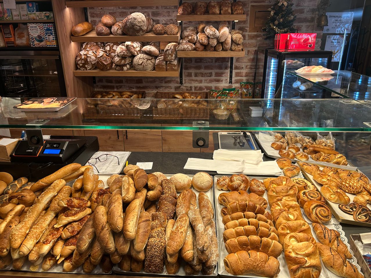 Eine Bäckerei-Vitrine mit verschiedenen Broten und Gebäck, darunter Laibe, Brötchen und Grissini, auf Tabletts mit Glasfronten und Regalen, die mit mehr Brot gefüllt sind.