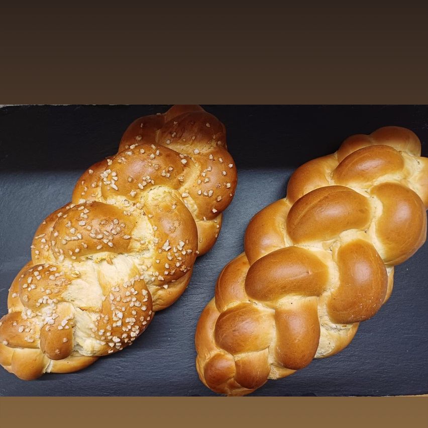 Zwei Challahbrote auf einer dunklen Oberfläche. Eines hat Sesamkörner obenauf.