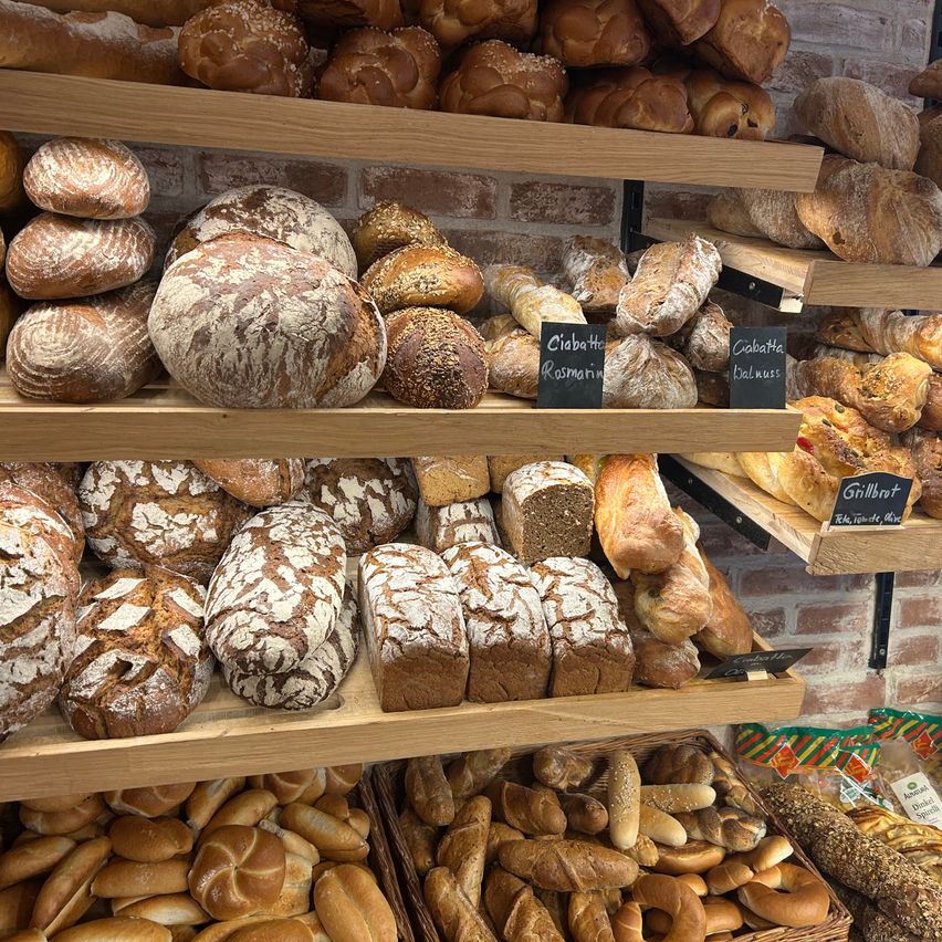 Eine Bäckerei-Auslage mit verschiedenen Brotsorten auf Holzregalen. Jedes Brot hat eine andere Textur und Belag. Einige Schilder zeigen die Namen der Brote.