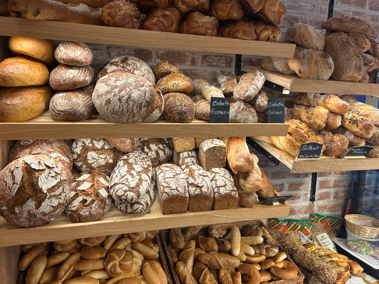 Eine Bäckerei-Auslage mit verschiedenen Brotsorten auf Holzregalen. Jedes Brot hat eine andere Textur und Belag. Einige Schilder zeigen die Namen der Brote.