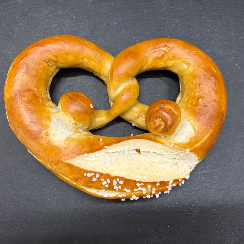 Ein herzförmiger Brezen mit abgebissenem Teil und Sesamkörnern liegt auf einer schwarzen Oberfläche.