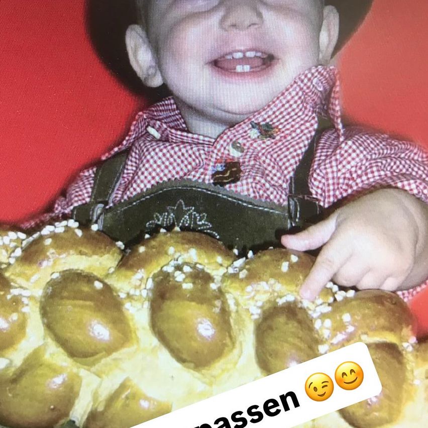Ein Kleinkind hält ein gebackenes Brot mit Sesamkörnern, trägt ein rotes Hemd und einen Hut. Er lächelt und scheint für ein Foto zu posieren.