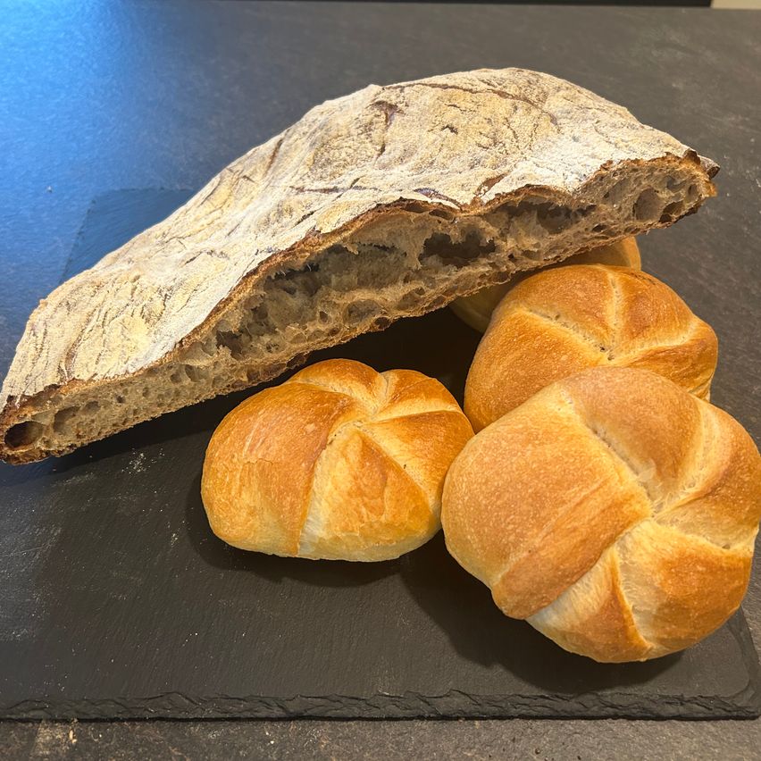 Ein Brotlaib mit einem geschnittenen Teil und drei Brötchen auf einer schwarzen Oberfläche. Das Brot ist goldbraun mit einer knusprigen Kruste.