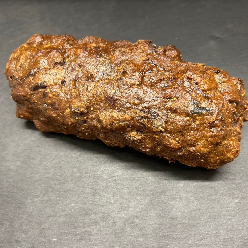 Eine Nahaufnahme eines langen, dunkelbraunen, unregelmäßig geformten Gegenstands auf einer Metalloberfläche. Es scheint sich um eine Art gekochtes Fleisch zu handeln, möglicherweise Wurst, mit einer knusprigen Außenschicht.