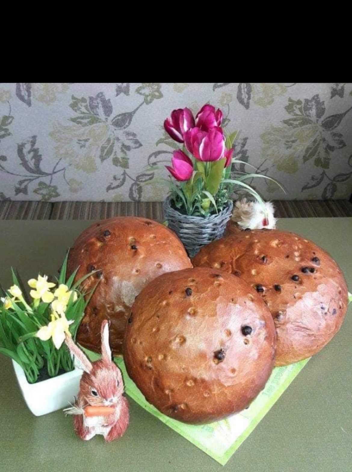 Drei kugelförmige Brote sind mit einer Vase mit rosa Tulpen und einer gelben Narzissenpflanze ausgestellt. Eine kleine Keramikhasenfigur hält eine Karotte.