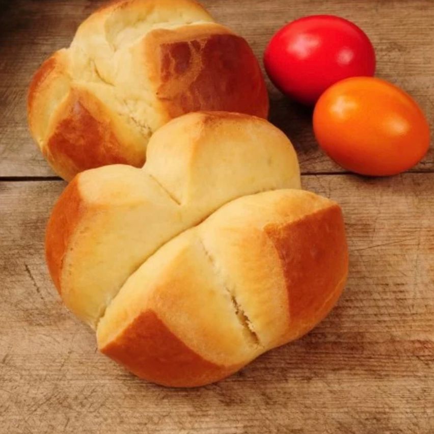 Zwei herzförmige Brötchen mit knuspriger Kruste liegen auf einer Holzoberfläche. Daneben befinden sich zwei bunte Eier, eines rot und eines orange.