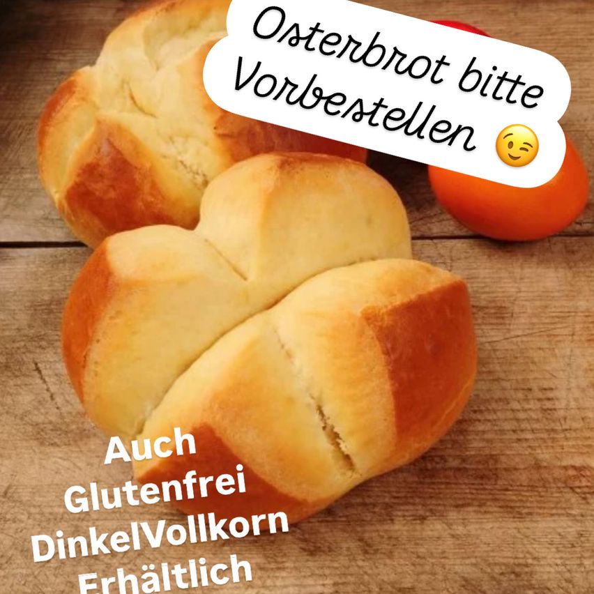 Zwei goldene Osterbrote auf einem Holztisch mit einer Sprechblase, die 'Osterbrot bitte vorbestellen' sagt. Eine Orange ist neben den Brötchen. Der Text enthält auch 'Auch Glutenfrei DinkelVollkorn Erhältlich'.