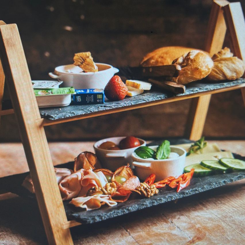 Ein Holztablett zeigt eine Auswahl an Lebensmitteln wie Brot, Käse, geschnittenem Fleisch, Tomaten und Gurken auf einer Holzoberfläche.