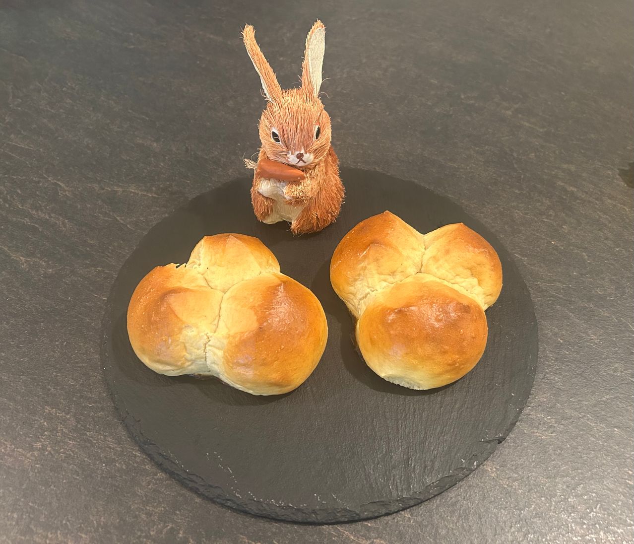 Ein kleines Kaninchen, das einen Keks hält, sitzt neben zwei Brötchen auf einem schwarzen Teller. Die Brötchen sind braun mit einer goldenen Kruste. Der Hintergrund ist grau.