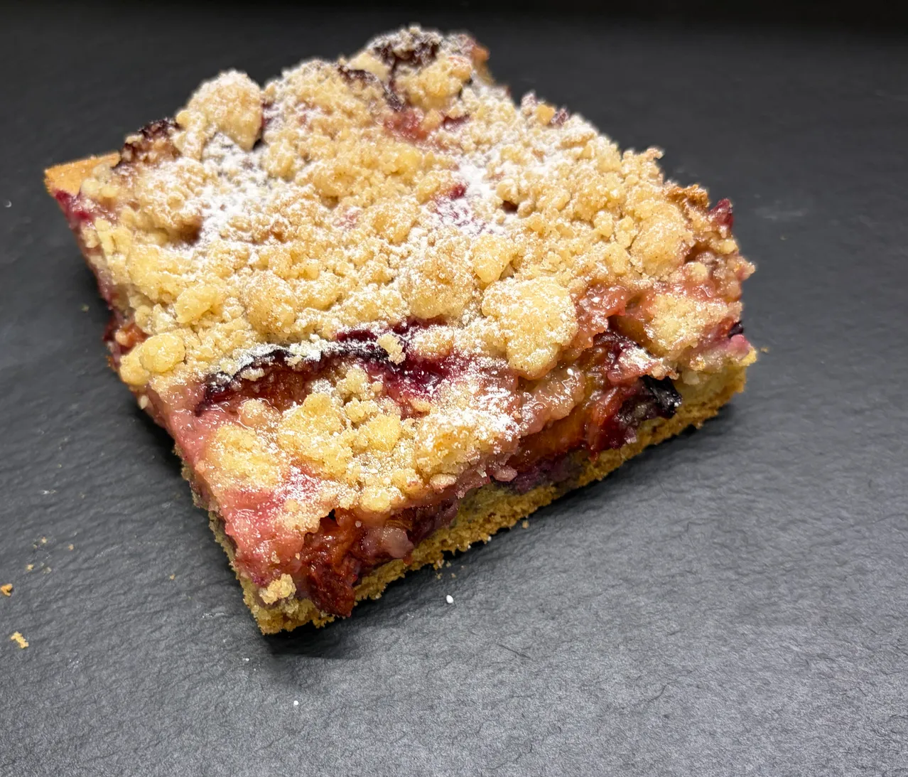 Ein quadratisches Stück Obst-Crumble-Kuchen mit knuspriger Kruste und Puderzucker obenauf.