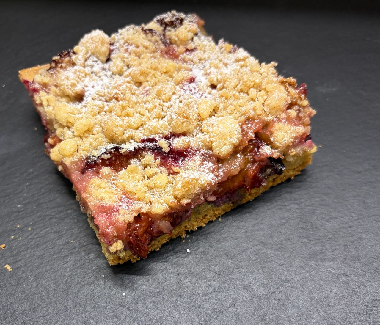 Ein quadratisches Stück Obst-Crumble-Kuchen mit knuspriger Kruste und Puderzucker obenauf.