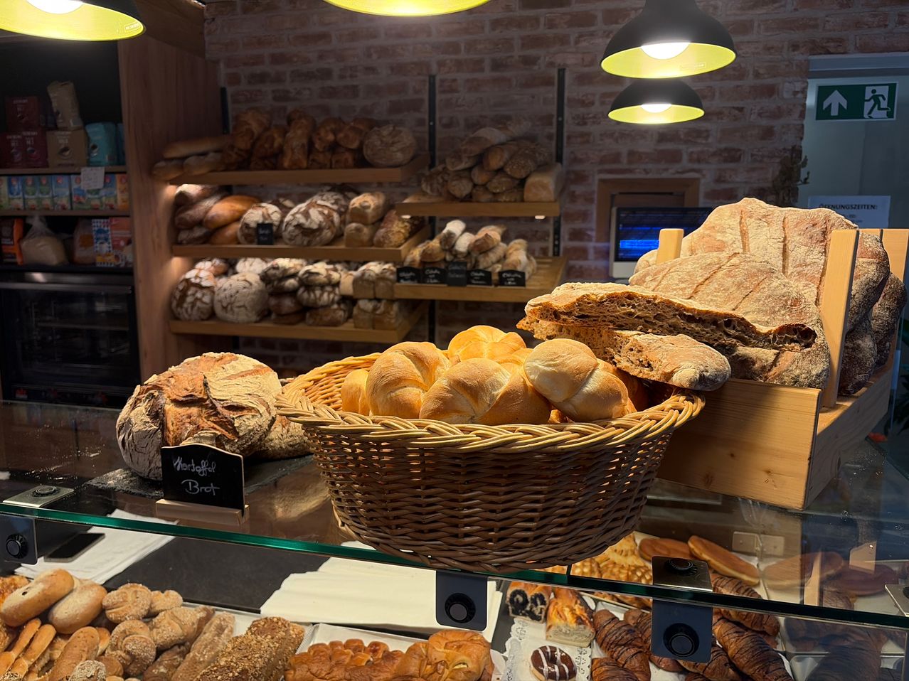 Eine Bäckerei-Auslage mit verschiedenen Broten und Gebäck in einem Korb und auf Regalen, beleuchtet von warmen gelben Lichtern.