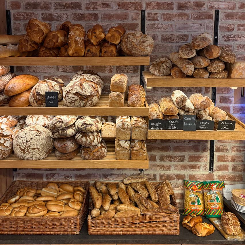 Eine Bäckerei-Auslage mit verschiedenen Brotsorten auf Holzregalen an einer Ziegelwand. Es sind auch Körbe mit Brot vorhanden.