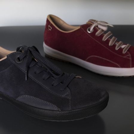 Zwei Sneaker, einer schwarz und einer burgunderrot, liegen nebeneinander auf einer dunklen Oberfläche. Der burgunderrote Sneaker hat eine weiße Sohle und dekorative Schnürsenkel.